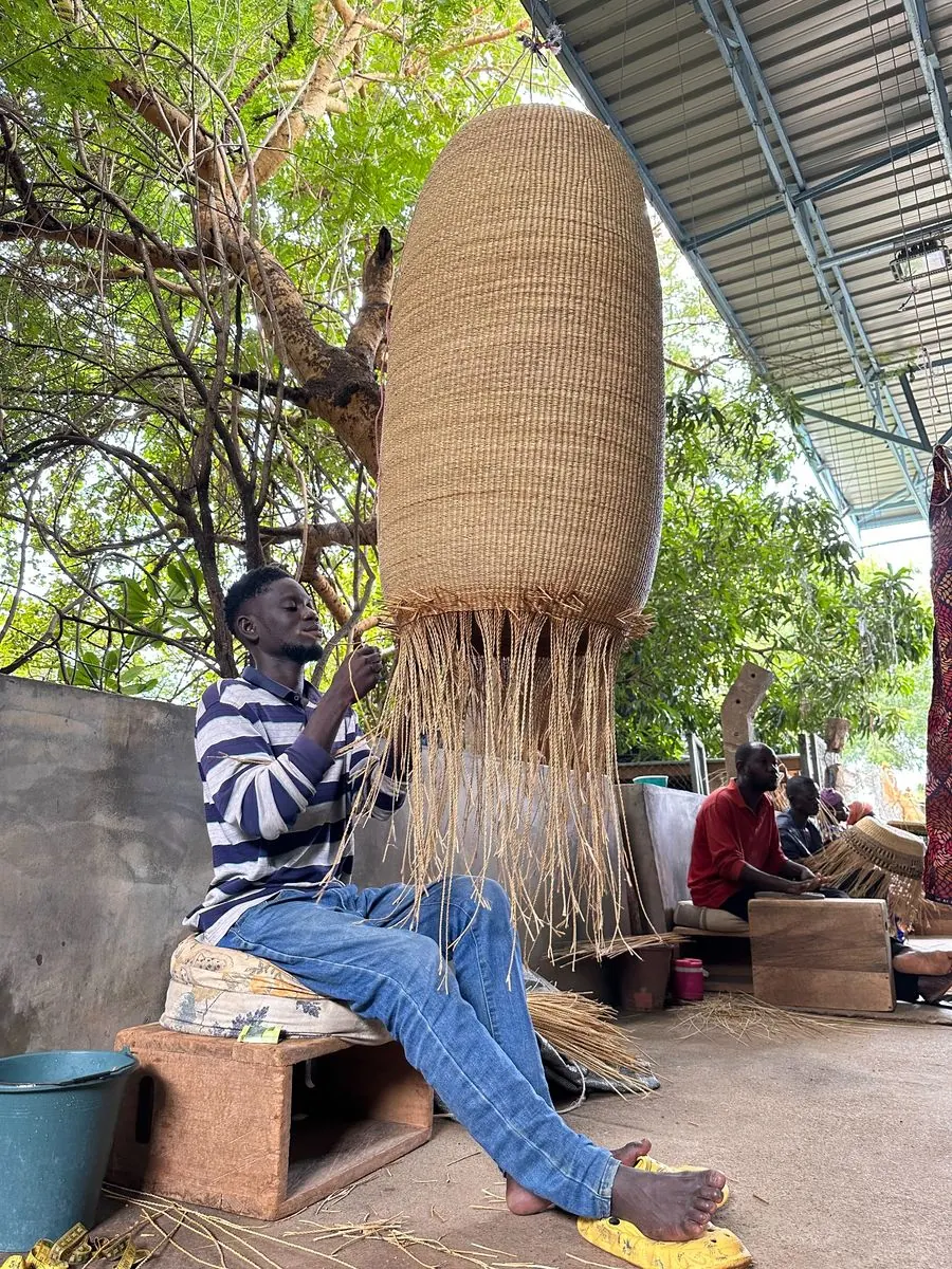 A tall natural straw basket hanging upside down while it is woven